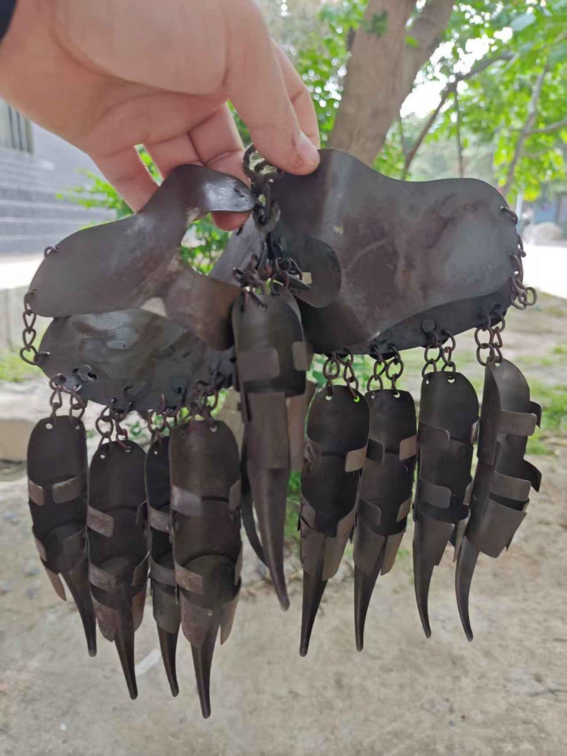 Pu&ograve; includere: Un elemento decorativo in metallo marrone scuro con una serie di elementi appesi e appuntiti. Il pezzo &egrave; tenuto in mano e sembra essere fatto di metallo martellato, con una catena che collega i vari componenti. Il design ha un'estetica unica, forse tribale.