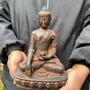 Może przedstawiać: Brązowa statua Buddy w pozycji siedzącej medytacji. Posąg ma skomplikowane detale, w tym wzorzyste szaty i spokojny wyraz twarzy. Posąg stoi na ozdobnej podstawie i ma zwietrzały wygląd.