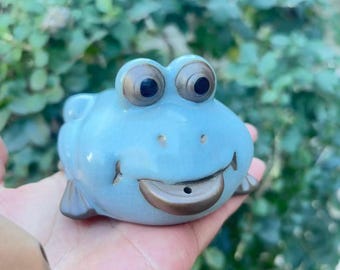 May include: A light blue ceramic frog figurine with large, expressive eyes and a smiling mouth. The frog has a smooth, glossy finish and is resting on a hand. The figurine is a decorative item.
