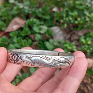 May include: A silver bangle bracelet featuring an engraved design of flowers and fish. The bracelet is held in a hand, with a blurred green background of foliage. The bracelet appears to be made of metal.