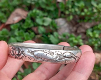 May include: A silver-colored bangle bracelet with an engraved floral and fish design. The bracelet is held in a hand, with a blurred green background of foliage. The bracelet appears to be made of metal.