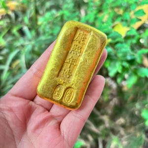 May include: A gold-coloured rectangular ingot with Chinese characters embossed on the surface. The ingot is held in a hand against a blurred green background. The characters are in a vertical format.