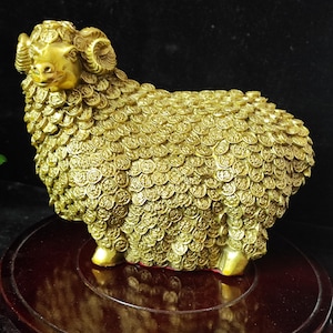 Puede incluir: Figurilla dorada de un carnero con intrincados detalles en forma de monedas que cubren su cuerpo. El carnero tiene cuernos curvados y está sobre una base de madera oscura. Objeto decorativo.