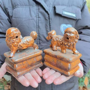 May include: Two ceramic dog figurines in a reddish-brown hue. Each dog is perched atop a rectangular base. The dogs have detailed fur and stand in a playful pose. The bases have a textured, aged appearance.