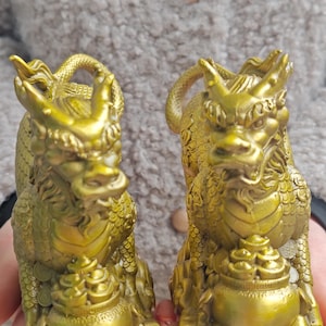 May include: Two gold-colored dragon figurines, each holding a round object that resembles a pot of gold. The dragons are facing forward with their mouths open, and their bodies are covered in intricate details.