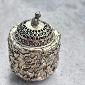 Könnte beinhalten: Ein detaillierter Räuchergefäß aus Messing mit einer kleinen Affenfigur oben. Der Deckel hat ein Wabenmuster und der Körper ist mit aufwendigen Reliefschnitzereien verziert. Der Brenner hat eine Vintage-Ästhetik.