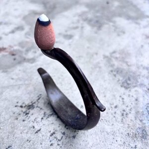 May include: A dark metal incense holder with a curved design, supporting a brown incense cone with a white tip. The holder is set against a textured, light-colored surface, creating a contrast between the dark metal and the lighter background.