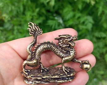Puede incluir: Una detallada figura de dragón de bronce con escamas intrincadas, una cola fluida y una expresión feroz. El dragón está posado sobre una base texturizada, sostenido en una mano contra un fondo verde borroso.