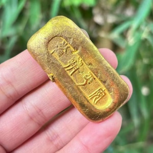 May include: A rectangular gold ingot with embossed Chinese characters. The ingot has a slightly textured surface and a rounded top. The background is blurred, suggesting an outdoor setting.