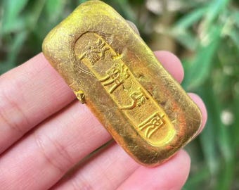May include: A rectangular gold ingot with embossed Chinese characters. The ingot has a slightly textured surface and a rounded top. The background is blurred, suggesting an outdoor setting.