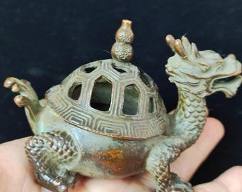 May include: A bronze incense burner in the shape of a turtle with a dragon head. The turtle's shell has a geometric pattern with cut-out shapes. The burner has a small, decorative top and is held in a hand.