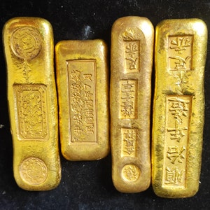 May include: Four gold bars with embossed Chinese characters and designs. The bars vary in size and shape, with rectangular and oblong forms. The gold has a bright, reflective sheen, suggesting a high purity. The bars are likely historical or collectible items.