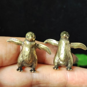 May include: Two small, bronze-colored penguin figurines with outstretched wings. The penguins have detailed features, including beaks, eyes, and feet. The figurines are standing on a surface, with a dark background.