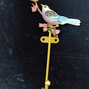May include: A decorative wall hook featuring a colorful bird perched on a branch with pink gemstone accents. The bird has blue, green, and purple feathers. The hook is gold-colored and has a rounded bottom.