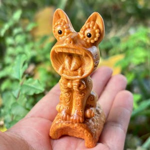 Puede incluir: Una detallada talla de madera de una criatura mítica con la boca abierta, orejas grandes y patrones intrincados. La escultura es de color marrón claro cálido y parece estar tallada a mano. La criatura está sentada sobre una base texturizada.