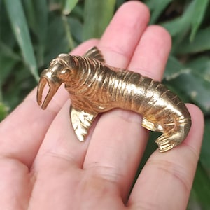 May include: A detailed, golden walrus figurine. The sculpture features a textured surface, visible tusks, and flipper details. The walrus is posed in a relaxed position, showcasing its distinctive features. The figurine is held in a hand against a blurred green background.