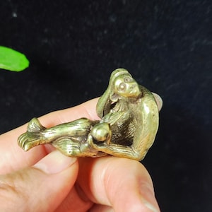 Puede incluir: Pequeña figura de mono de color latón en una pose relajada. El mono está tumbado, sosteniendo una pequeña esfera, con la cabeza apoyada en la mano. La figura tiene detalles y un acabado pulido.
