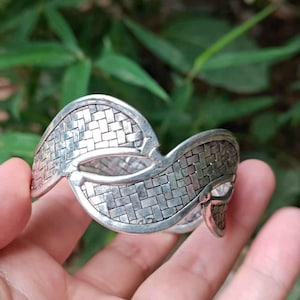 May include: A silver cuff bracelet with a woven basket weave pattern. The bracelet has an open design with curved edges and a textured surface. The background is blurred green foliage.