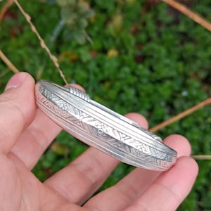 Könnte beinhalten: Silberfarbenes Armband mit aufwendigen floralen und geometrischen Mustern. Das Armband hat eine ovale Form und scheint aus Metall zu sein. Das Design umfasst detaillierte Gravuren auf der Oberfläche, die einen Vintage- oder Antik-Stil zeigen.