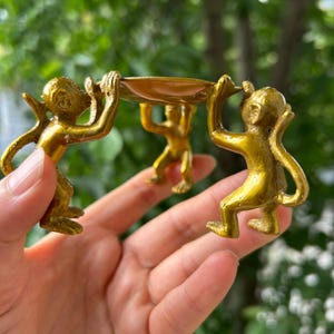 Può includere: Un vassoio decorativo color oro sostenuto da tre statuette di scimmia. Le scimmie sono accovacciate, che sostengono il vassoio con le braccia. Il vassoio è di colore oro chiaro e lo sfondo è verde sfocato.