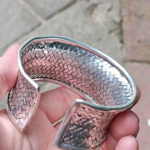 May include: A silver cuff bracelet with a woven basket weave design. The bracelet has a curved shape and an open design. The metal has a shiny, reflective surface. The bracelet is a piece of jewellery.