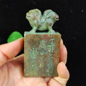 May include: A weathered bronze-colored seal with a double-headed animal figure on top. The rectangular base features engraved Chinese characters. The seal has a green patina, suggesting age. The background is dark.
