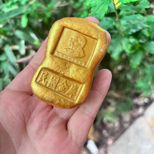 May include: A gold-coloured, rectangular ingot with embossed Chinese characters and a portrait. The ingot has a slightly irregular shape and is held in a hand against a background of green foliage. The text on the ingot is in Chinese.