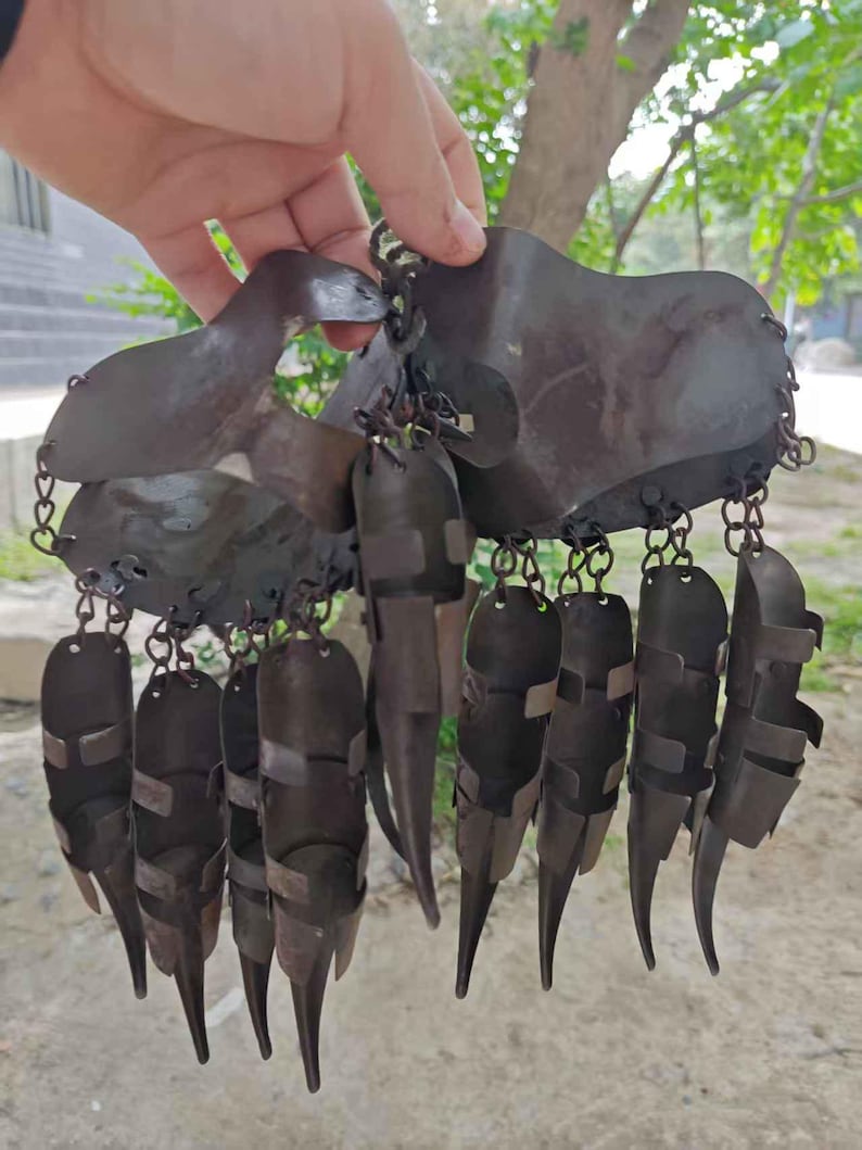 Pu&ograve; includere: Un elemento decorativo in metallo scuro con una serie di elementi appesi e appuntiti. Il pezzo &egrave; tenuto in mano e sembra essere fatto di un metallo scuro, forse invecchiato. Il design include una struttura centrale curva con molteplici forme allungate pendenti.