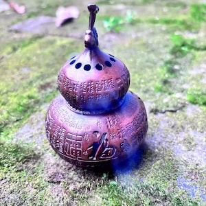 Könnte beinhalten: Ein bronzefarbener Räuchergefäß in Form einer Doppelgourde mit einem vogelartigen Aufsatz. Die Oberfläche ist mit chinesischen Schriftzeichen graviert. Der obere Teil hat kreisförmige Löcher. Auf grünem Moos platziert.