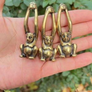 May include: Three small, brass-colored monkey figurines. Each monkey is in a seated position with its arms raised above its head. The figurines have a detailed, aged appearance, suggesting they are vintage or antique.