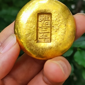 May include: A circular, gold-coloured ingot with embossed Chinese characters. The surface has a textured appearance, and the ingot is held in a hand against a blurred green background. The characters are in a rectangular frame.