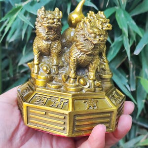 May include: A golden figurine featuring three mythical creatures with detailed features, standing on a hexagonal base with Chinese characters. The statue is held in a hand, with a green leafy background.