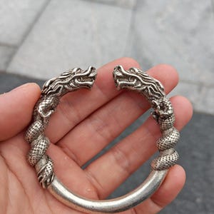 May include: Silver bracelet with two dragon heads facing outwards. The bracelet has a thick, curved band with detailed dragon scales and a textured, twisted design. The bracelet is held in a hand.