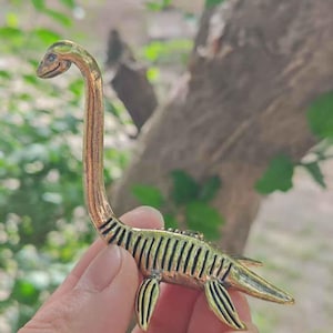 Peut inclure: Une figurine stylisée de couleur bronze représentant un plésiosaure, un reptile marin préhistorique. La figurine a un long cou, une petite tête et des appendices en forme de nageoires. Le corps est détaillé avec des marques nervurées.