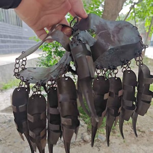 Pu&ograve; includere: Una scultura artigianale a forma di artiglio in metallo scuro. Il pezzo presenta un elemento centrale a forma di scudo collegato a pi&ugrave; segmenti a forma di artiglio tramite catene. Il metallo ha un aspetto invecchiato, che suggerisce un'estetica antica o rustica.
