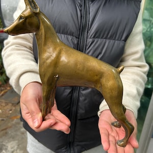May include: A bronze-colored dog sculpture, likely a Doberman Pinscher, is held in front of a person. The sculpture is detailed, showing the dog's slender body, pointed ears, and alert posture. The bronze has a slightly aged patina.