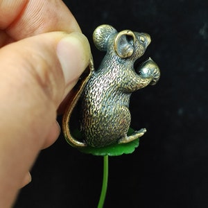 Può includere: Statuetta di topo color bronzo che tiene una noce, appollaiata su una foglia verde. Il topo presenta dettagli precisi e una lunga coda. La statuetta è posta su uno sfondo scuro.