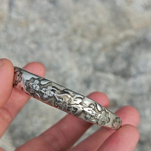 May include: A silver-colored bangle bracelet with a floral design. The bracelet is engraved with detailed flowers and leaves. The metal has a slightly tarnished appearance, giving it a vintage look. The bracelet is held in a hand.