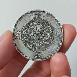 May include: A silver coin with a detailed dragon design at the centre, encircled by Chinese characters. The coin has a textured edge and is held in a hand, highlighting its intricate details and historical significance.