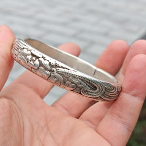 Puede incluir: Un brazalete plateado con intrincados diseños florales y de volutas. El brazalete tiene una banda ancha y una forma ligeramente curvada. Los grabados detallados cubren toda la superficie, creando una apariencia texturizada. El brazalete se sostiene en una mano.