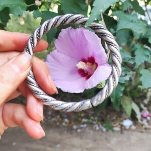 Könnte beinhalten: Ein silberfarbenes, gedrehtes Metallarmband. Das Armband wird vor einer hellvioletten Blume mit einem dunkelroten Zentrum gehalten. Der Hintergrund zeigt grüne Blätter und Laub.