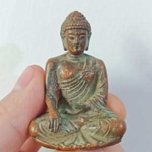 Könnte beinhalten: Kleine Buddha-Statue aus Bronze in sitzender Meditationshaltung. Die Statue hat ein verwittertes, gealtertes Aussehen mit Braun- und Grüntönen. Die Hände des Buddha ruhen im Schoß, und der Kopf hat ein detailliertes, strukturiertes Design.