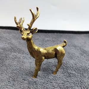 May include: A small, detailed brass deer figurine. The deer is standing with its head up and antlers prominent. The figurine has a golden hue and is set against a gray background. It is a decorative item.