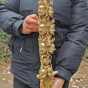 Puede incluir: Una daga ritual dorada y ornamentada con múltiples rostros y una figura de Buda en la parte superior. La daga tiene un extremo puntiagudo y se sostiene verticalmente. Los rostros son detallados y el diseño es intrincado, lo que sugiere un propósito religioso o ceremonial.