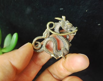 Puede incluir: Un amuleto de metal dorado en forma de dragón con detalles intrincados. El dragón está enroscado con la cabeza levantada y la cola envuelta alrededor de su cuerpo. El amuleto tiene un bucle en la parte superior para sujetarlo a un collar o pulsera.