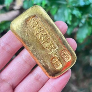May include: A rectangular gold bar with embossed Chinese characters. The bar has a slightly rounded top and is held in a hand. The gold has a shiny, reflective surface.