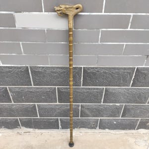 May include: A gold-coloured walking cane featuring a dragon head handle. The cane has a long, slender shaft with vertical ridges. The dragon head is intricately detailed, with a curved neck and open mouth. The cane's tip is black.