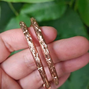 May include: Two gold-colored hoop earrings with a starburst pattern. The earrings are held in a hand against a blurred green background. The earrings have a shiny, metallic finish and a simple, elegant design.