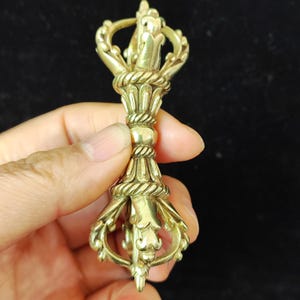 May include: A detailed, gold-coloured vajra, a ritual object, is held in a hand. The vajra features intricate designs, including a central knot and ornate ends. The object has a symmetrical shape and appears to be made of metal.