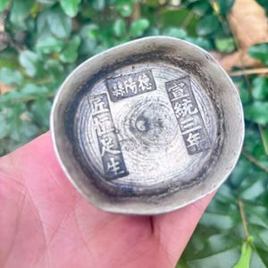 May include: A small, silver-coloured metal bowl with Chinese characters stamped inside. The bowl has a slightly irregular shape and a textured surface. The characters are arranged in rectangular blocks. The bowl is held in a hand against a background of green leaves.